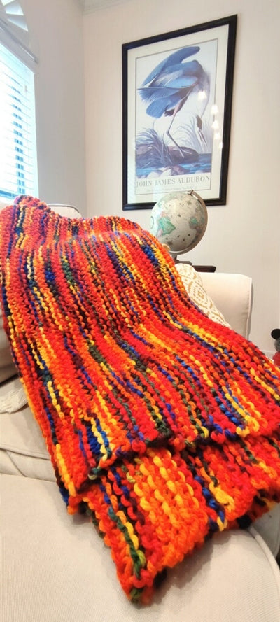 The blanket is primarily red with yellow, orange, green, and blue woven in. The blanket sits on top of pillows on a tan couch with a globe in the background a painting on the wall.