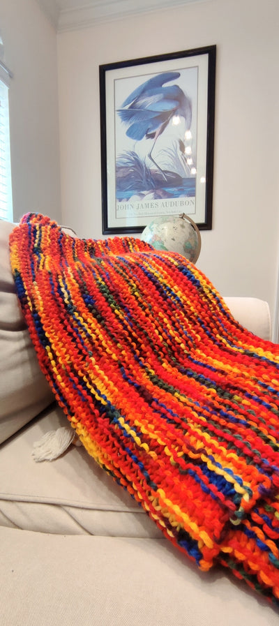 The blanket is primarily red with yellow, orange, green, and blue woven in. The blanket sits on top of pillows on a tan couch with a globe in the background a painting on the wall.