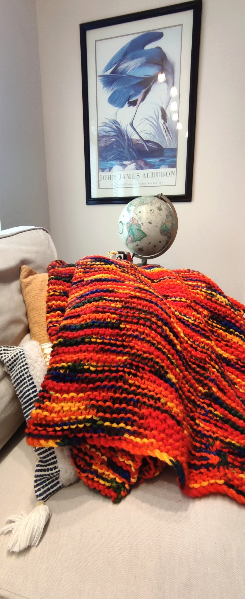 The blanket is primarily red with yellow, orange, green, and blue woven in. It sits on a tan couch with a globe in the background.