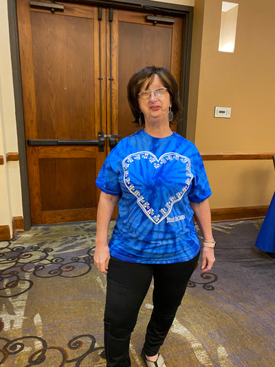 A woman stands in front of brown double doors. She is wearing a light blue, blue, and white tie-dye shirt features the Braille ABCs lining the inside of a hand-drawn heart. It's printed in our puff ink so that it is tactile! 
