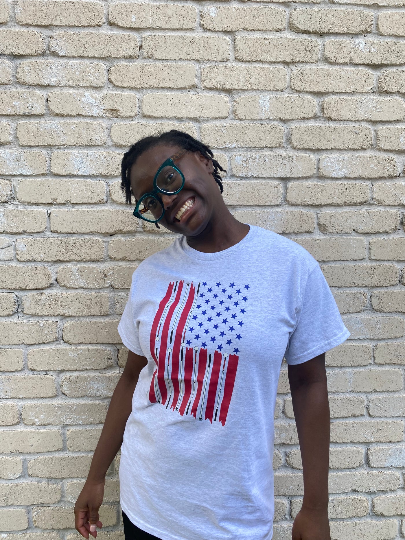 Cyndi is wearing a light grey t-shirt with a large print of a flag hanging downward is on the chest. The red stripes resemble hand drawn stripes. White canes take the place of white stripes. The 50 stars are blue with no background.