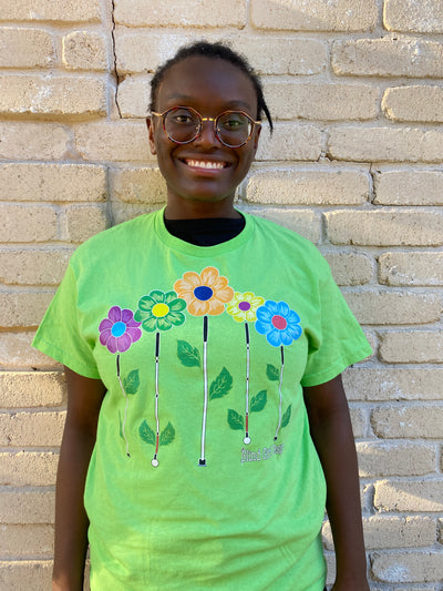 Cyndi stands against a cream colored brick wall and is smiling. This lovely lime t-shirt has 5 graphic design flowers - purple with a sky blue middle, green with a yellow middle, tangerine with a navy middle, yellow with a purple middle, and sky blue with a fuchsia middle - with blind canes as stems to make a lovely garden! The canes are even sprouting darling little leaves.