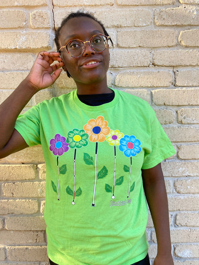 Cyndi stands against a cream colored brick wall and is smiling. This lovely lime t-shirt has 5 graphic design flowers - purple with a sky blue middle, green with a yellow middle, tangerine with a navy middle, yellow with a purple middle, and sky blue with a fuchsia middle - with blind canes as stems to make a lovely garden! The canes are even sprouting darling little leaves.