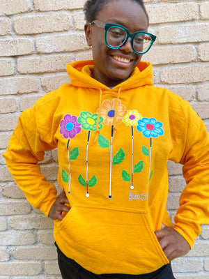 Cyndi in golden yellow flower garden hoodie against a cream colored brick wall