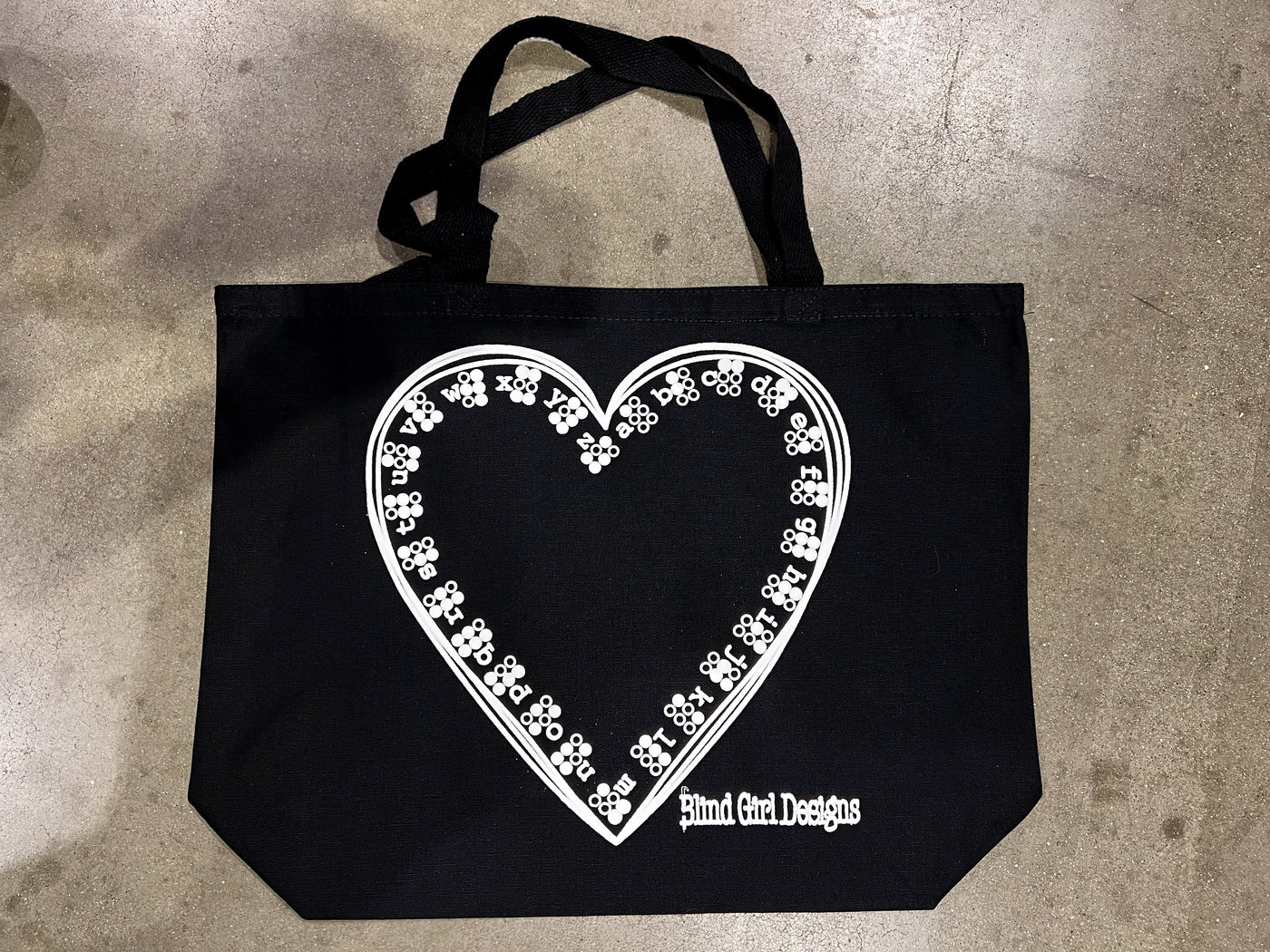 A black canvas tote with the Braille heart print, a hand drawn 12 x 12 heart, inside the perimeter of the heart, starting at the top, is the braille alphabet a through Z with adjacent letters.