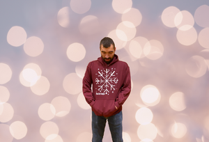 Uless, a 6 foot tall male with dark brown short hair, stands with a dark red sweatshirt on that has a snowflake made of three different white came styles. There are smaller snowflakes sprinkled around the large center image. His hands are in the front pocket and he's looking down.