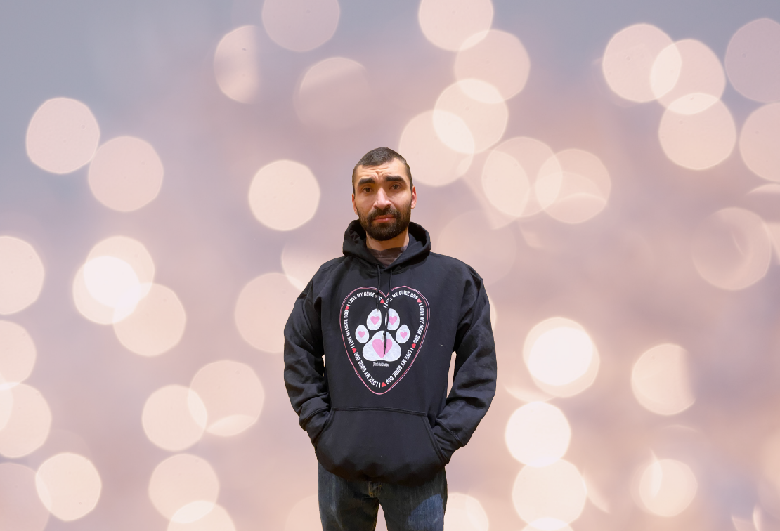 Uless, a 6 foot tall male with dark brown short hair, stands with his hands in the front pocket of a black hoodie. The hoodie as the I LOVE MY GUIDE DOG print on the front. 