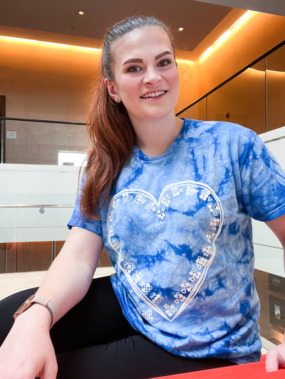 A girl sits on stairs and wears a light blue, blue, and white tie-dye shirt features the Braille ABCs lining the inside of a hand-drawn heart. It's printed in our puff ink so that it is tactile! 