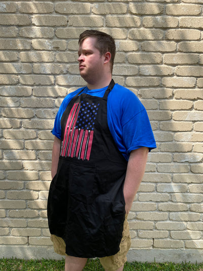 Robert, a tall white male, stands outside against a brick wall and wears a black apron with our American flag created by laying out a traditional American flag, the stars are blue, the stripes are red, but the white stripes are actually white canes.