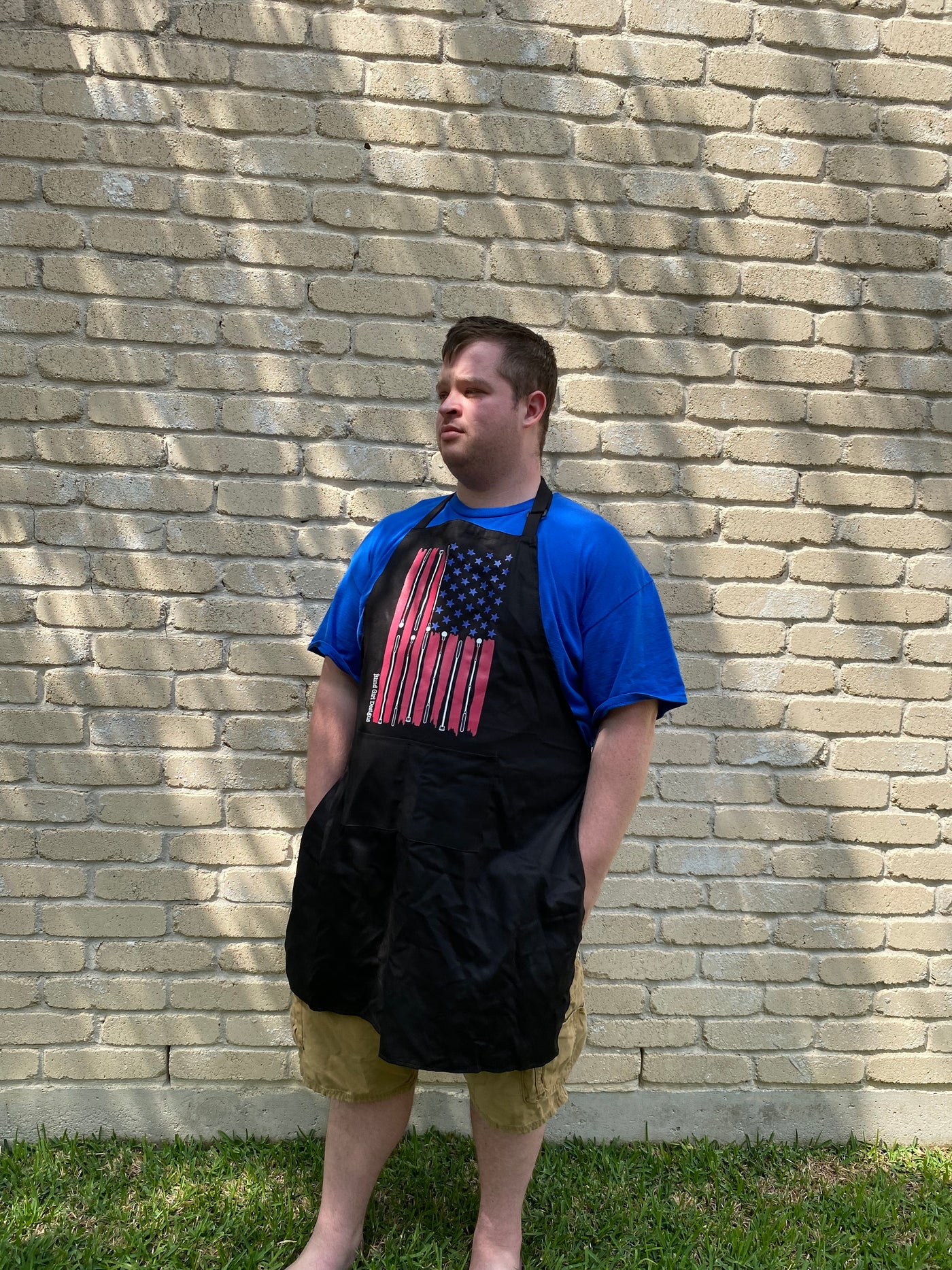 Robert, a tall white male, stands outside against a brick wall and wears a black apron with our American flag created by laying out a traditional American flag, the stars are blue, the stripes are red, but the white stripes are actually white canes.