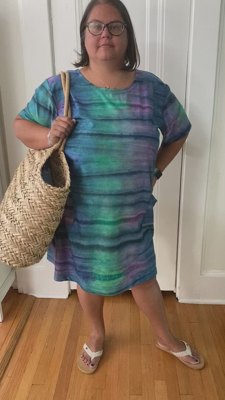 Model is facing the camera in front of a white door, wearing a dress of blue, purple, and green with navy lines that resembles the aurora borealis. She has paired the dress with white sandals and a tan, woven, oversized bag.
