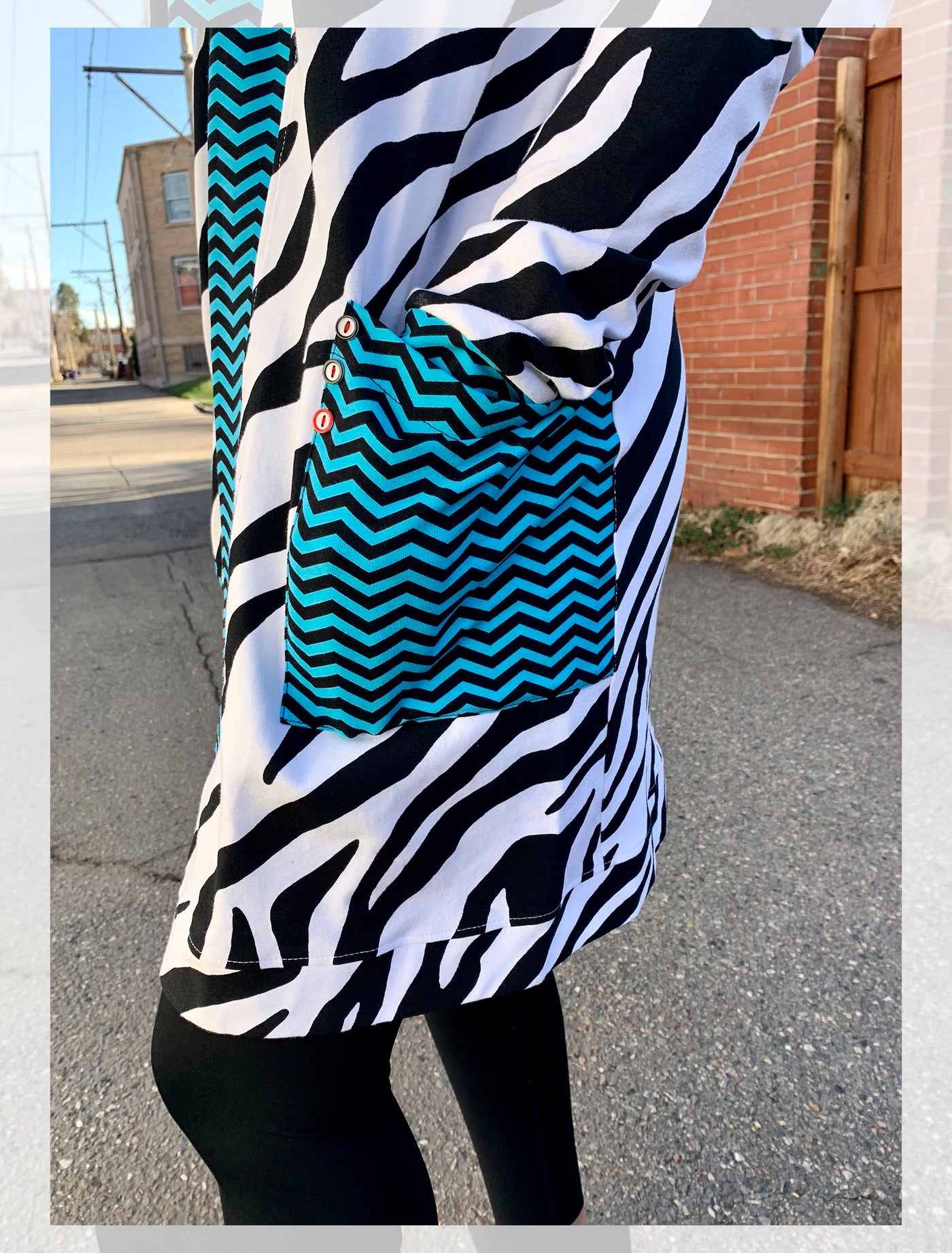Model stands outside wearing a knee length cardigan with electric blue and navy chevron edging and large, front pockets. The body of the cardigan is black and white zebra pattern. She has paired it with black leggings. The photo is a close up of her hand in the pocket.