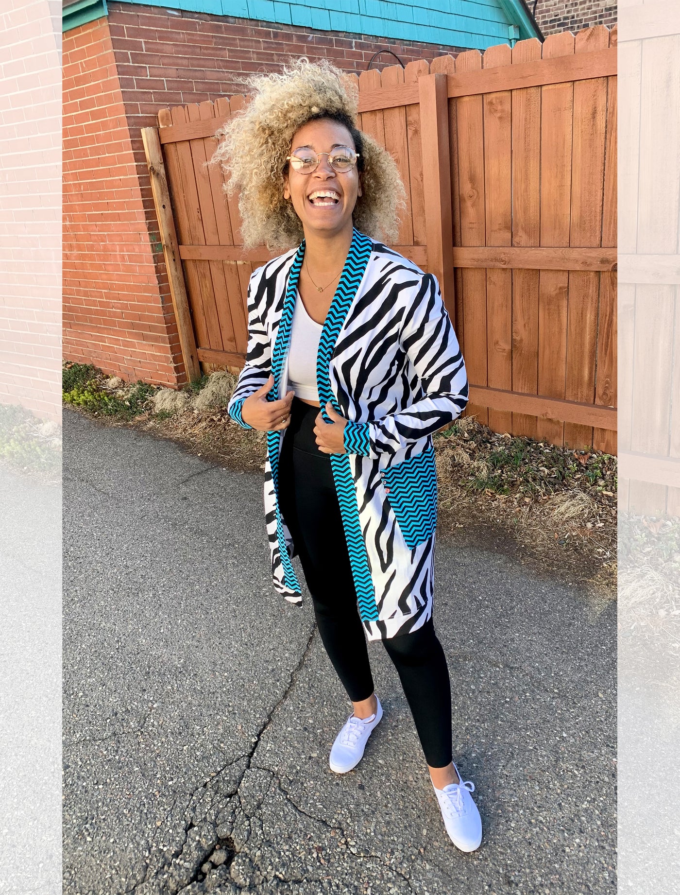 Model stands outside in front of a fence smiling as she wears a knee length cardigan with electric blue and navy chevron edging and large, front pockets. The body of the cardigan is black and white zebra pattern. She has paired it with black leggings, a white crop top and white sneakers.