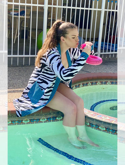 Model sits outside on the side of a pool wearing a knee length cardigan with electric blue and navy chevron edging and large, front pockets. The body of the cardigan is black and white zebra pattern. She is kissing a fake flamingo.