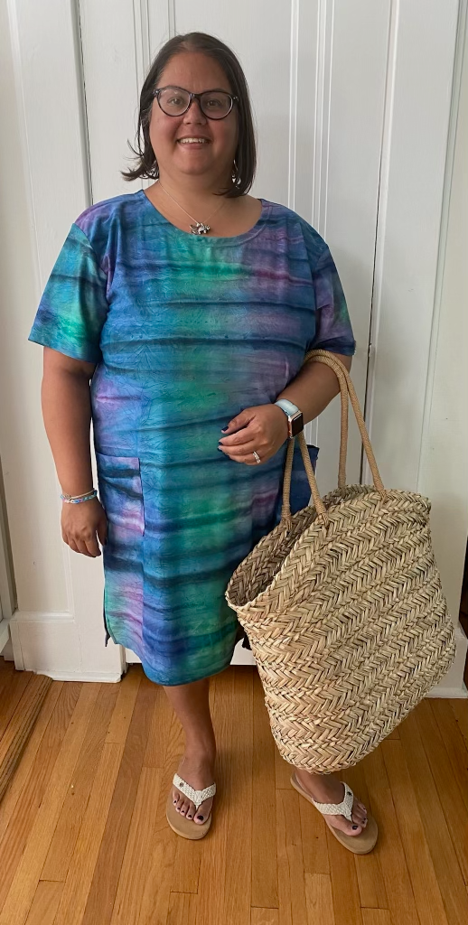Model is facing the camera in front of a white door, wearing a dress of blue, purple, and green with navy lines that resembles the aurora borealis. She has paired the dress with white sandals and a tan, woven, oversized bag.