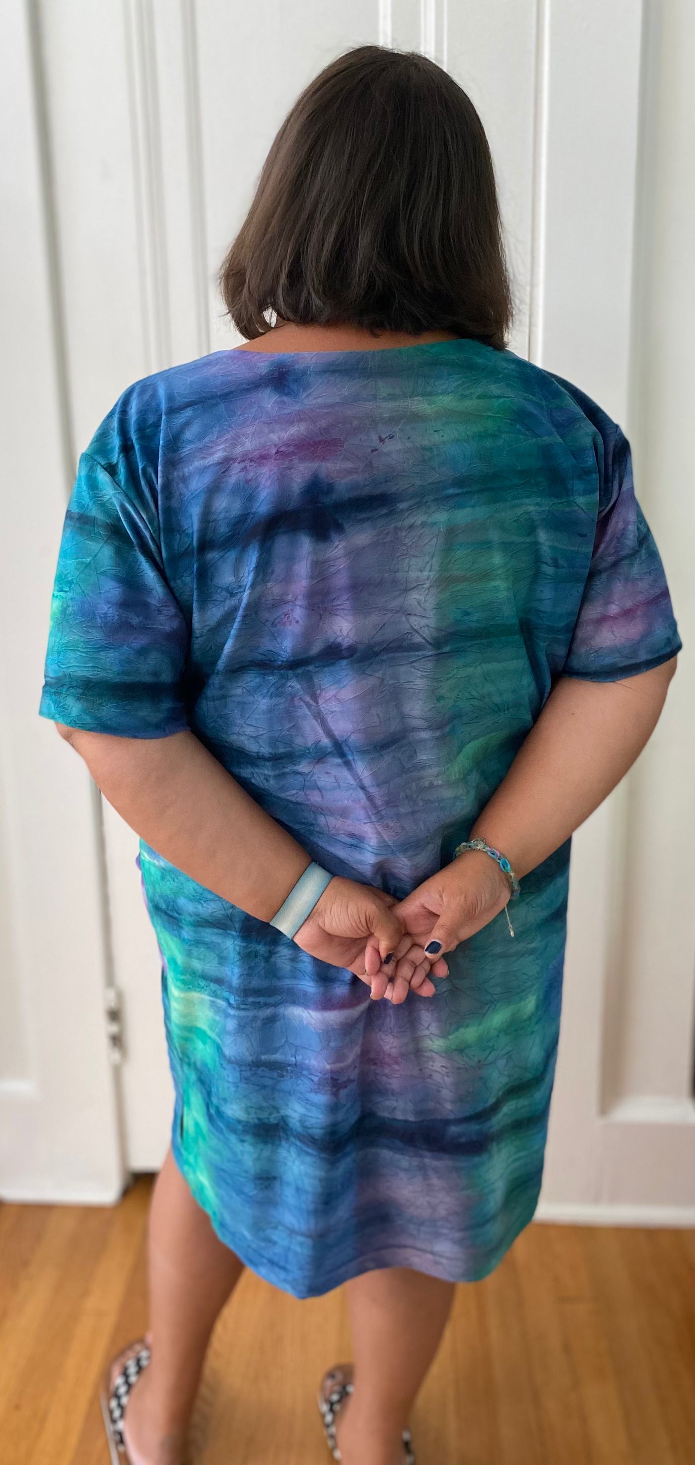 Model is facing away from the camera in front of a white door, wearing a dress of blue, purple, and green with navy lines that resembles the aurora borealis. Her arms are behind her back.