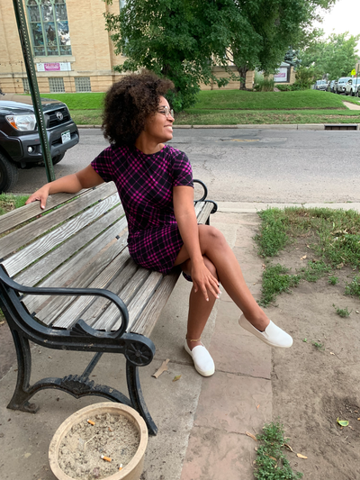 A young woman sits on a wooden bench in a neighborhood wearing a t-shirt dress. The dress is black and rich purple in a plaid pattern. She has paired it with white sneakers. She is smiling and wearing glasses.