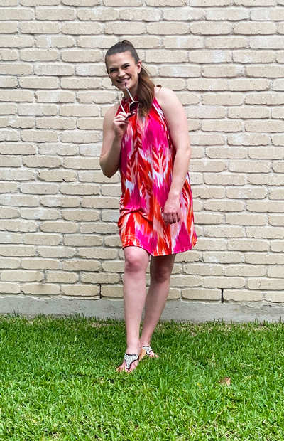 A young woman stands in front of a cream colored brick wall on green grass. She is wearing a loose, knee length dress that is bright pink, orange and white. She is smiling and holding onto her sunglasses.