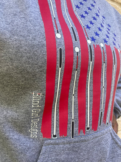 Cyndi stands against a cream colored brick wall with a light grey sweatshirt on that has the American flag has white canes in place of the white stripes between hand-drawn red stripes and perfectly placed 50 blue stars. 