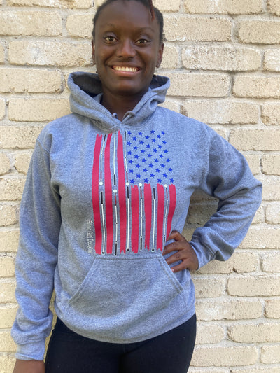 Cyndi stands against a cream colored brick wall with a light grey sweatshirt on that has the American flag has white canes in place of the white stripes between hand-drawn red stripes and perfectly placed 50 blue stars. 