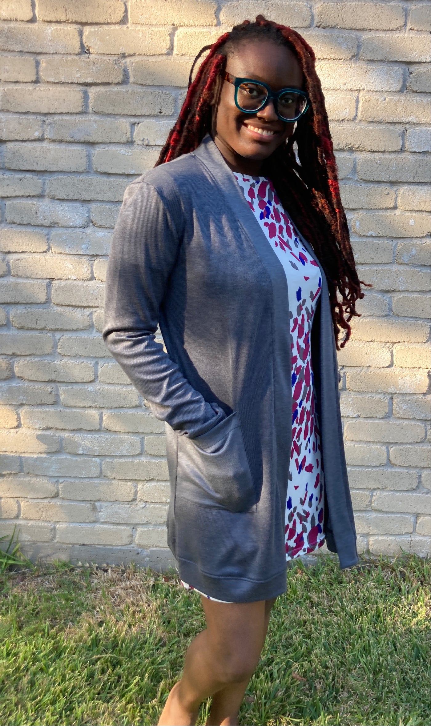 Model is wearing knee-length long sleeve and relaxed cardigan. The cardigan is open with no closures. The cardigan is heather grey on the inside and outside. She is posing outside against a cream colored brick wall.